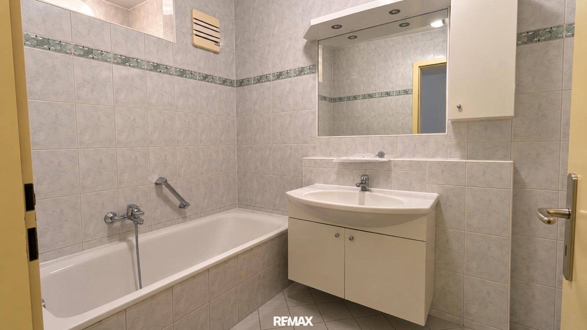 Helles Badezimmer mit hellgrauen Fliesen, Badewanne und Waschtisch mit Spiegelschrank.