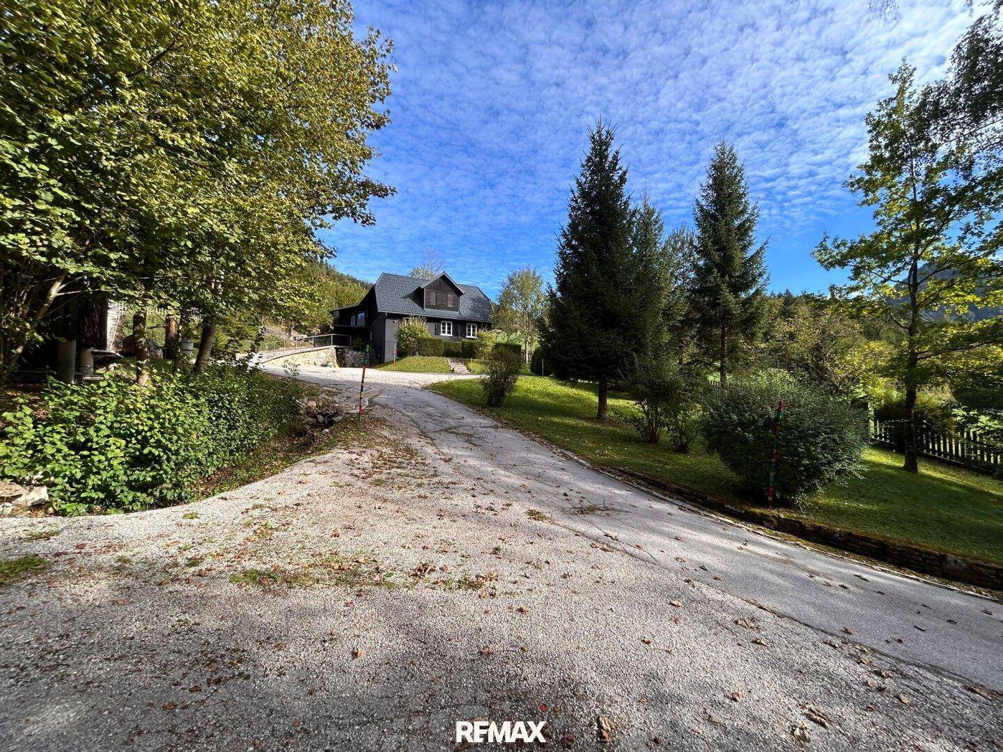 Geschotterte Auffahrt führt zum Haus, umgeben von Bäumen und mit Bergblick unter blauem Himmel.
