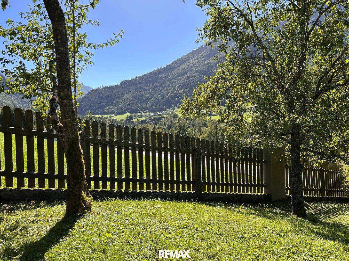 Holzzaun und grüne Wiese mit weitem Blick auf ein sonniges Tal und bewaldete Berge.