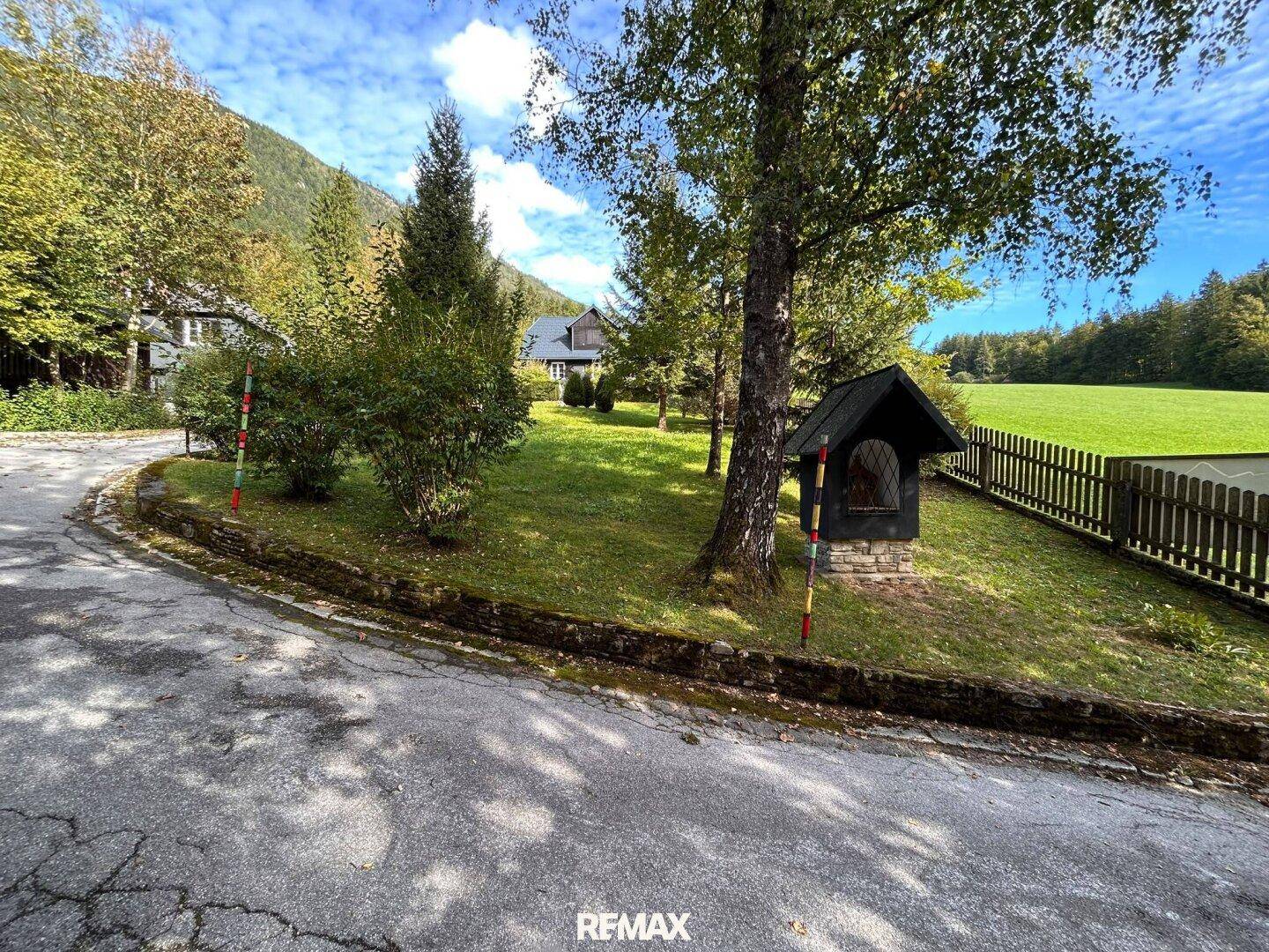 Ein Schotterweg führt durch einen Garten mit Bäumen und einem kleinen Schrein, umgeben von grünen Wiesen und Bergen.