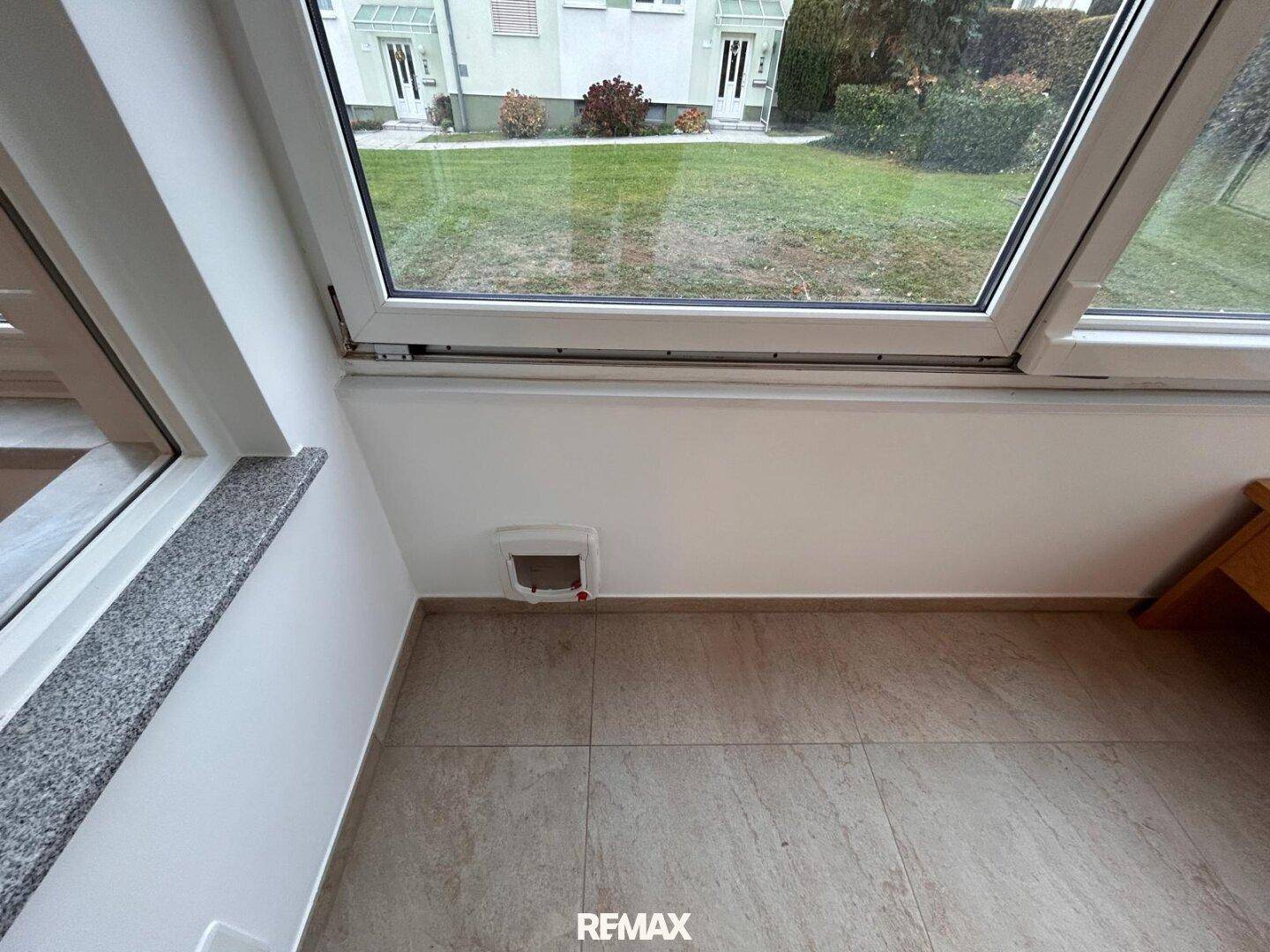 Detailansicht eines Fensters mit Katzenklappe und Blick auf den Vorgarten, gefliester Bodenbereich.