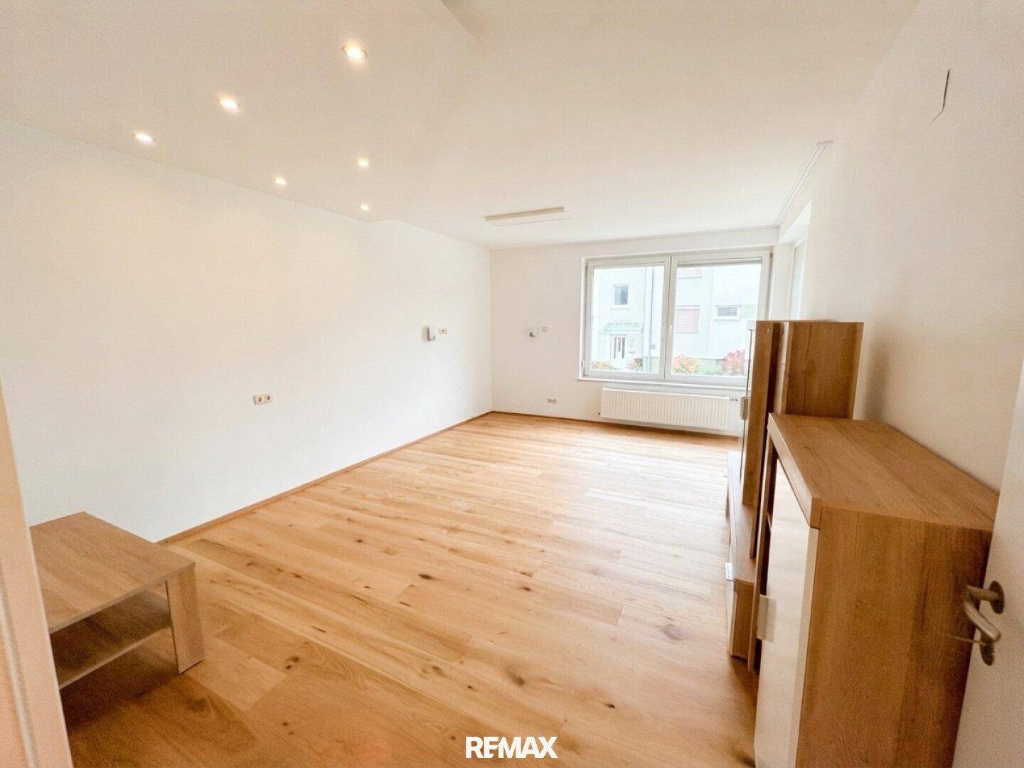 Geräumiges Wohnzimmer mit hellem Parkettboden und großen Fenstern, die viel Tageslicht hereinlassen.