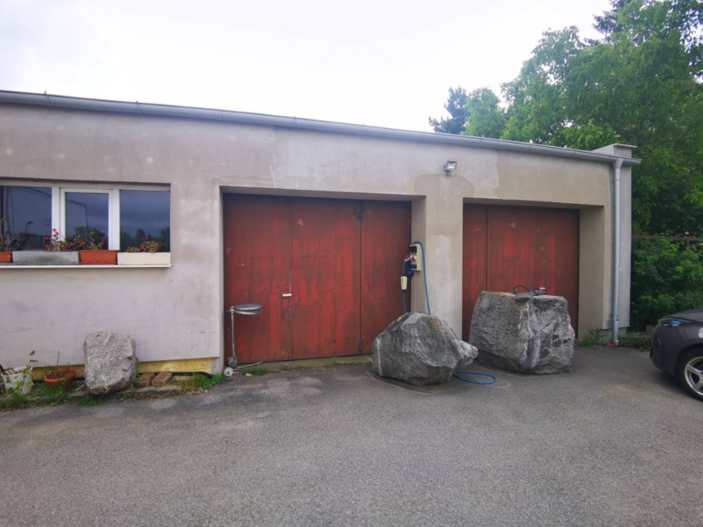 Außenansicht eines Gebäudes mit zwei roten Garagentoren und einem Fenster, davor große Steine.