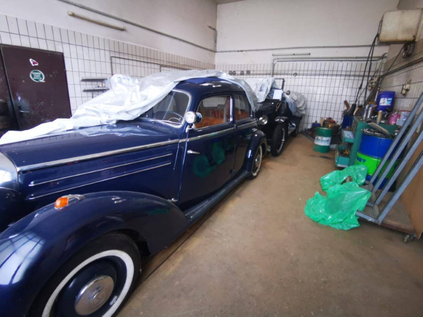 Innenansicht einer Garage oder Werkstatt mit gefliesten Wänden und mehreren Oldtimern unter Abdeckungen.