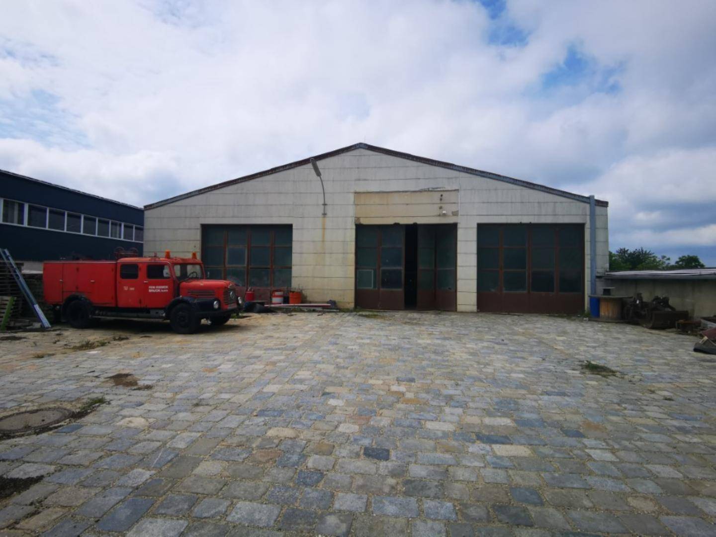 Große Industriehalle mit zwei breiten Toren und einem gepflasterten Hof, auf dem ein altes Feuerwehrauto steht.