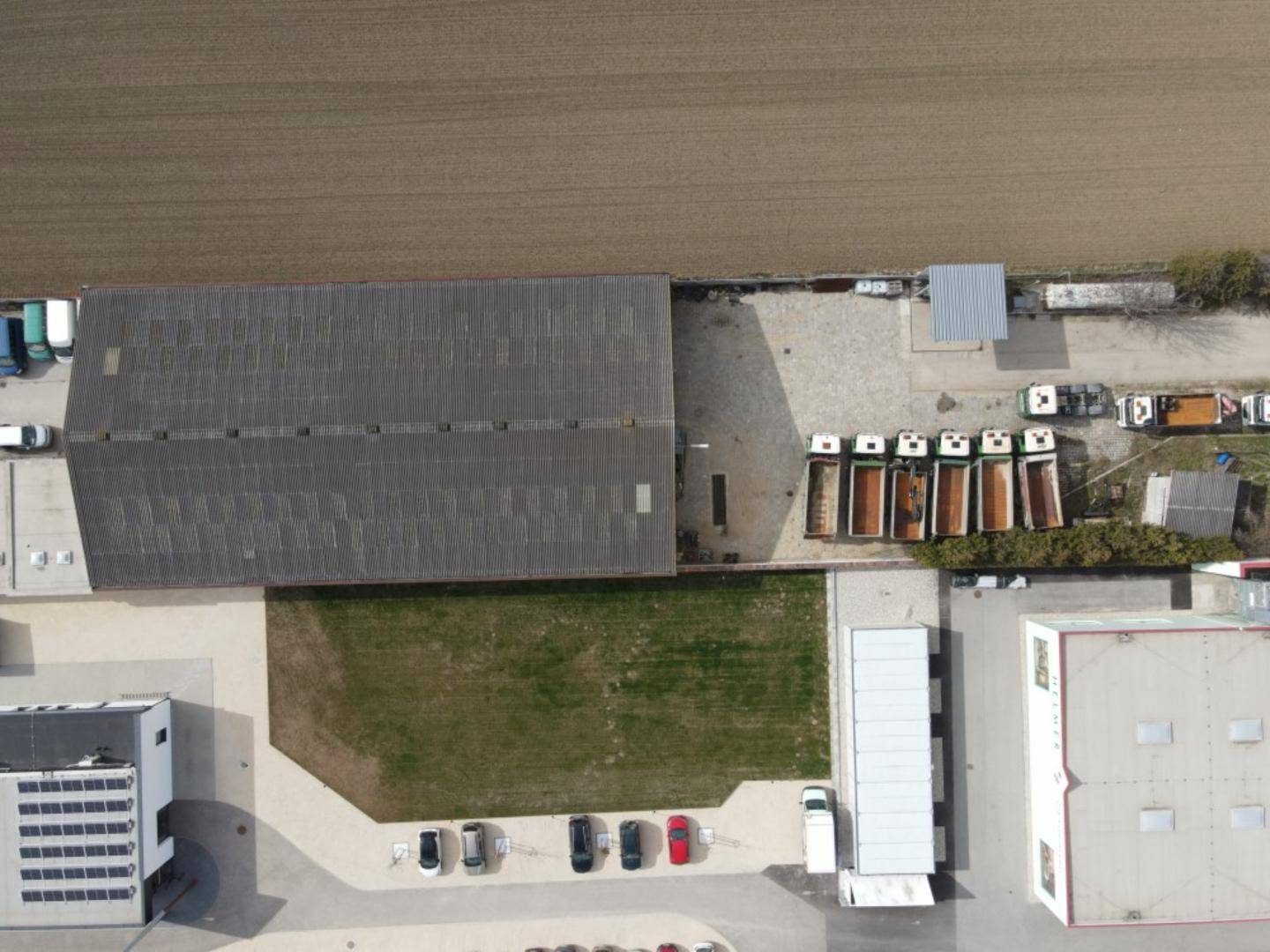 Luftaufnahme eines großen Betriebsgeländes mit Halle, Parkplätzen und angrenzenden Feldern.