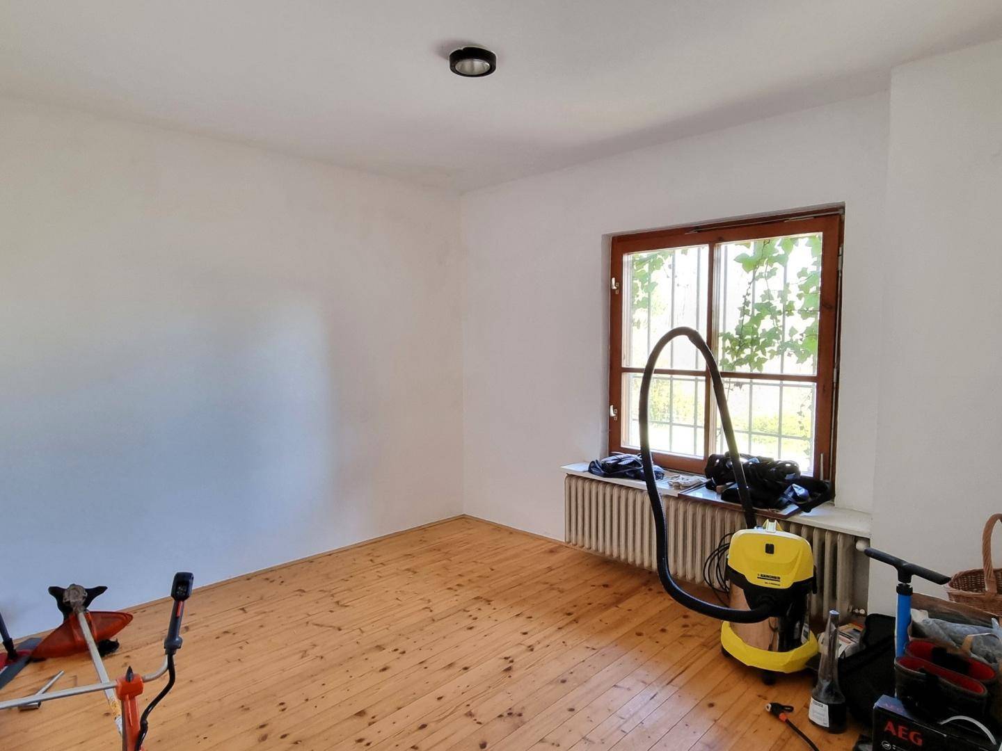Leerer Raum mit Holzboden, weiß gestrichenen Wänden und einem Fenster mit Gitter.