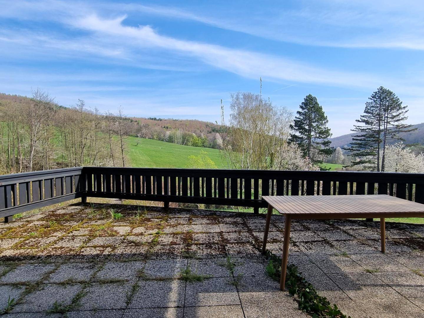 Große Terrasse mit Holztisch und weitem Blick über grüne Hügel und Bäume unter blauem Himmel.