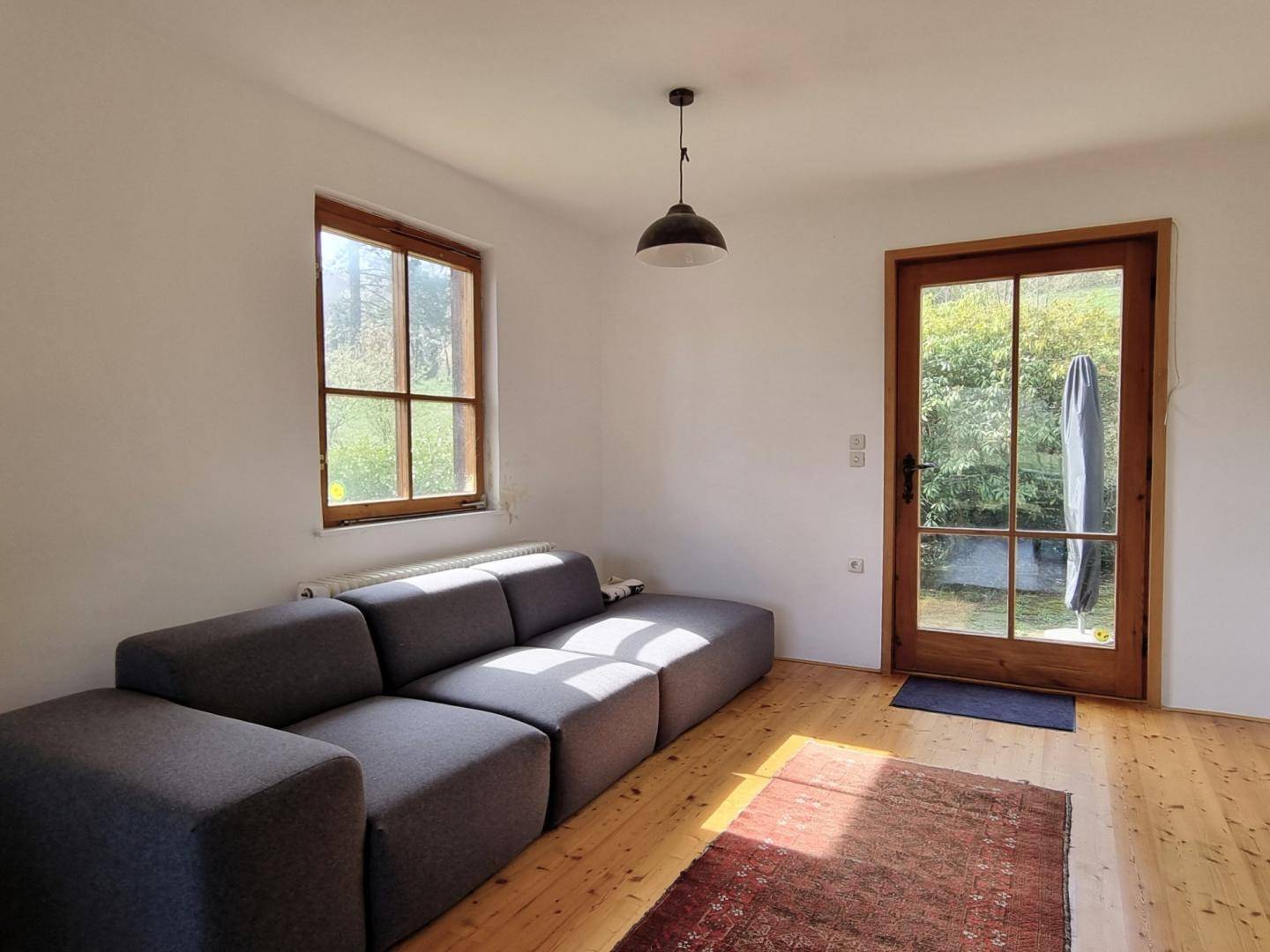 Gemütliches Wohnzimmer mit Holzboden, grauem Sofa und viel Tageslicht durch große Fenster.