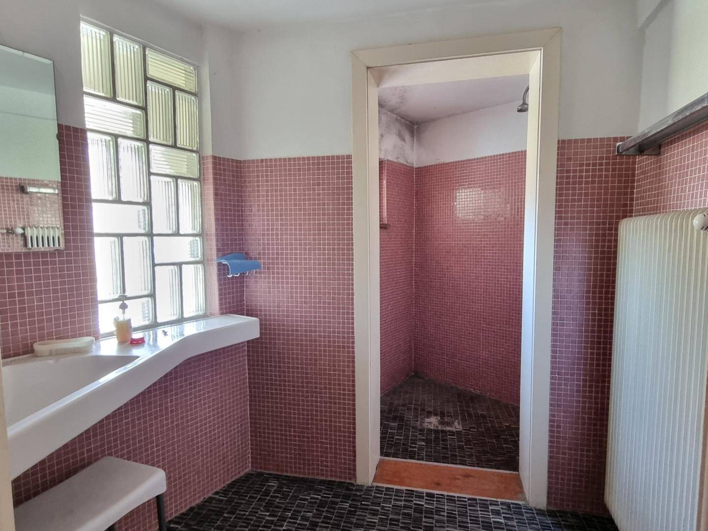 Badezimmer mit Duschbereich, Waschbecken und rosa Mosaikfliesen sowie einem Fenster aus Glasbausteinen.