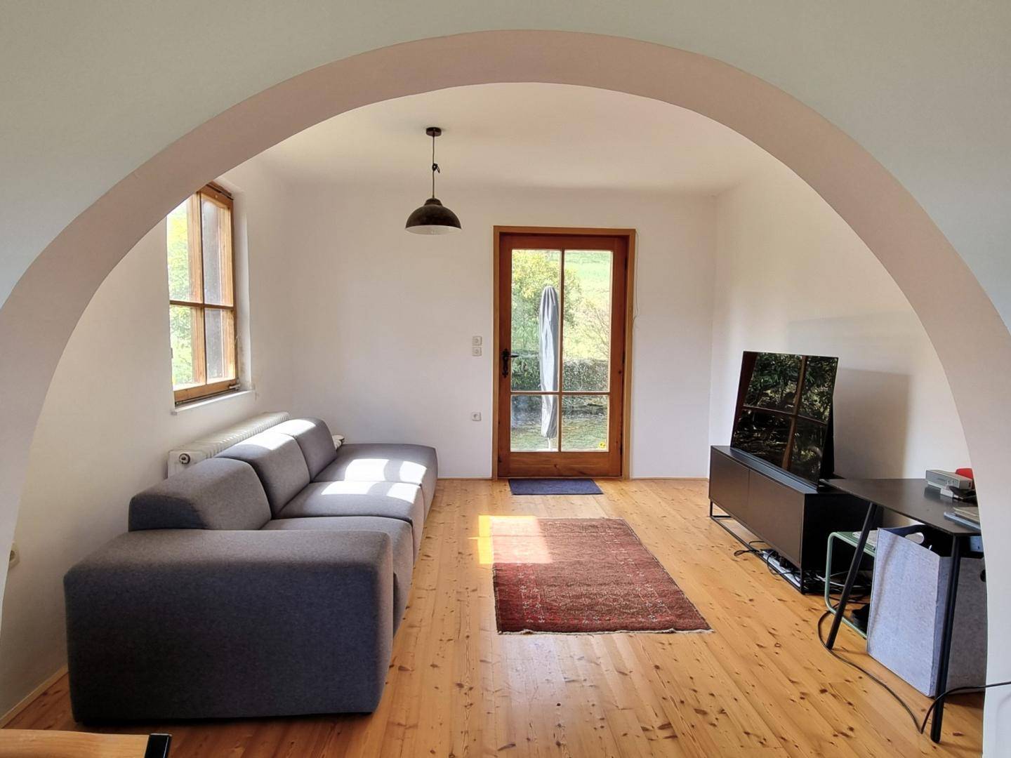 Helles Wohnzimmer mit Holzboden, modernem Sofa und Zugang zum Außenbereich durch eine Bogentür.