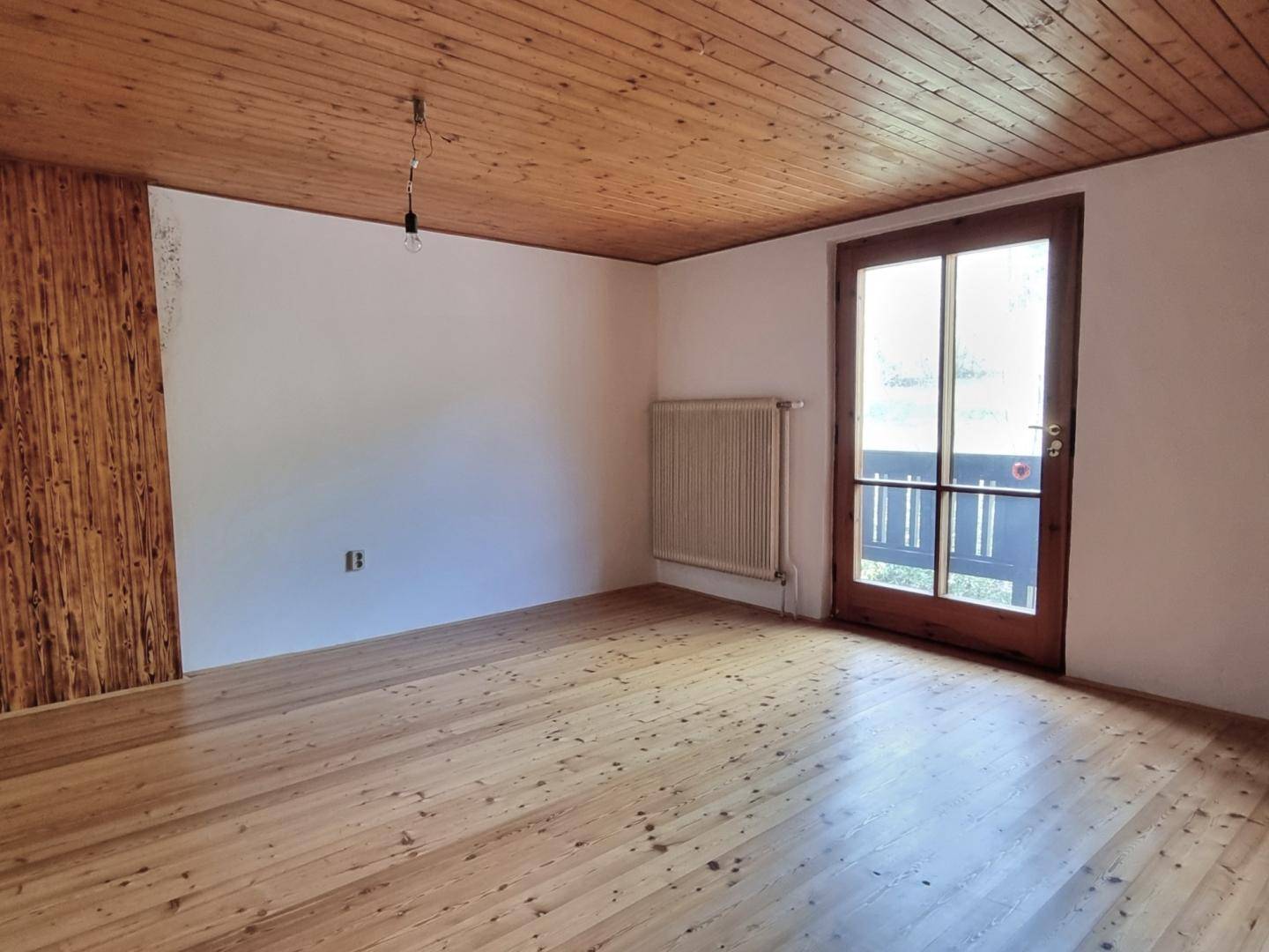 Heller Raum mit Holzboden, Holzdecke und Zugang zu einem Balkon mit Blick ins Grüne.