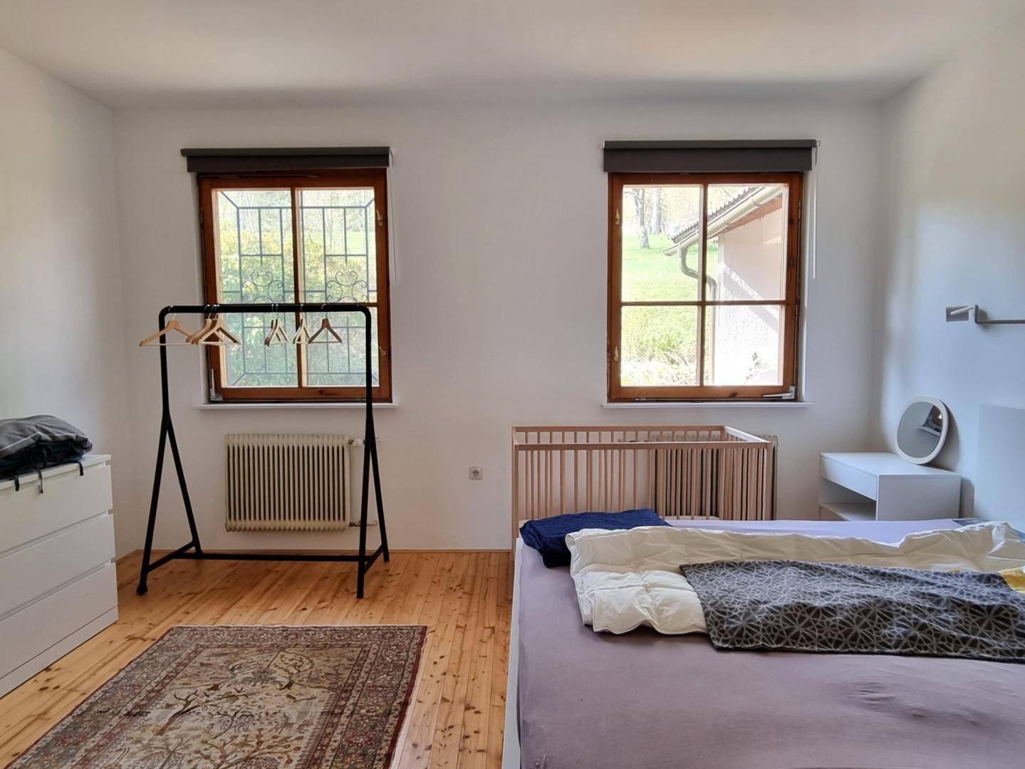 Helles Schlafzimmer mit Holzboden, zwei Fenstern und einem Kinderbett neben dem Doppelbett.