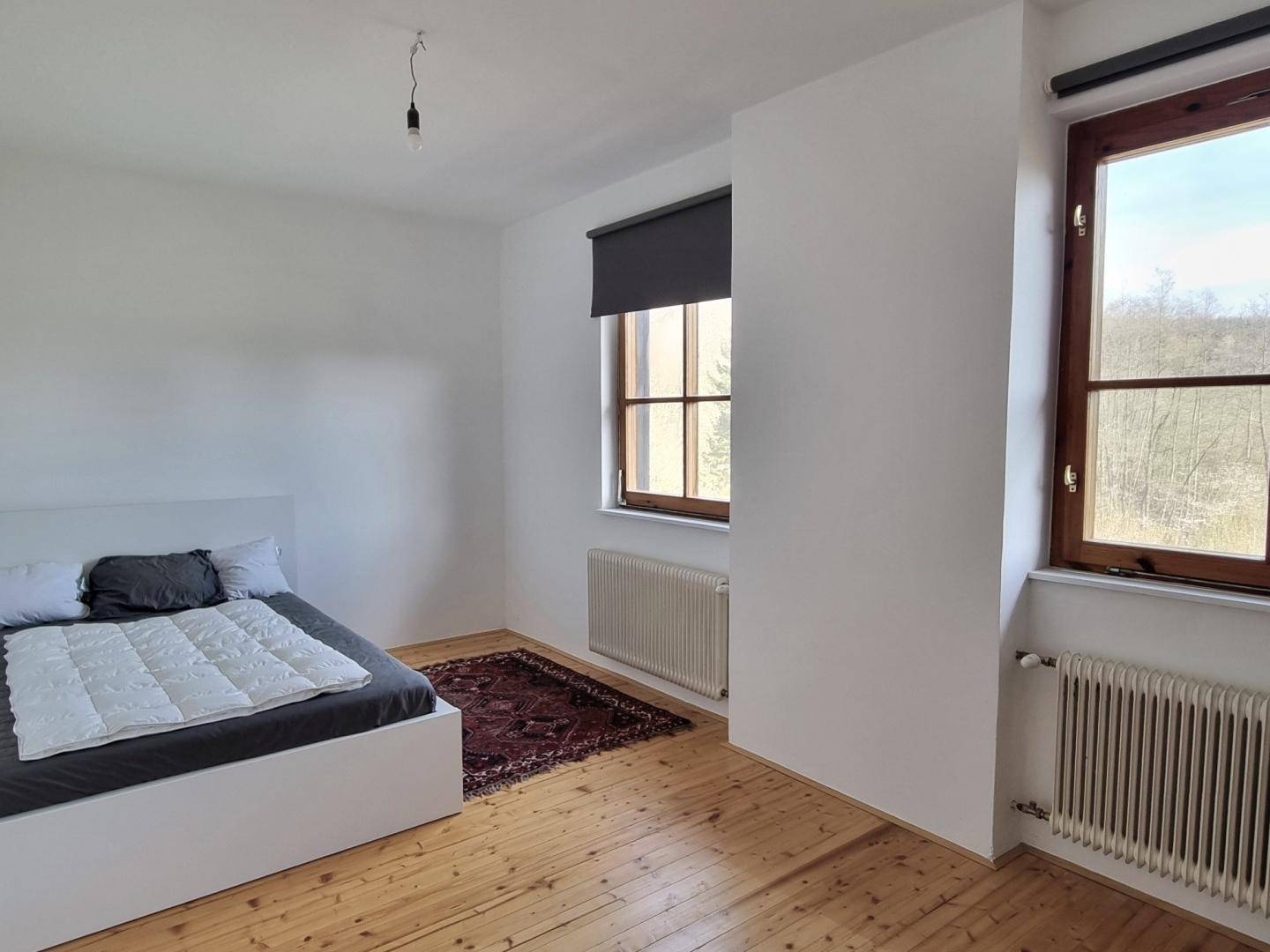 Schlafzimmer mit Holzboden, weißem Bett und zwei Fenstern, die viel Tageslicht hereinlassen.