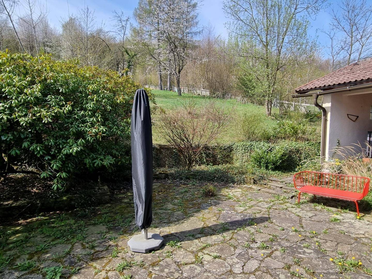 Gartenbereich mit gepflasterter Terrasse, Sonnenschirm und roter Bank, umgeben von viel Grün.