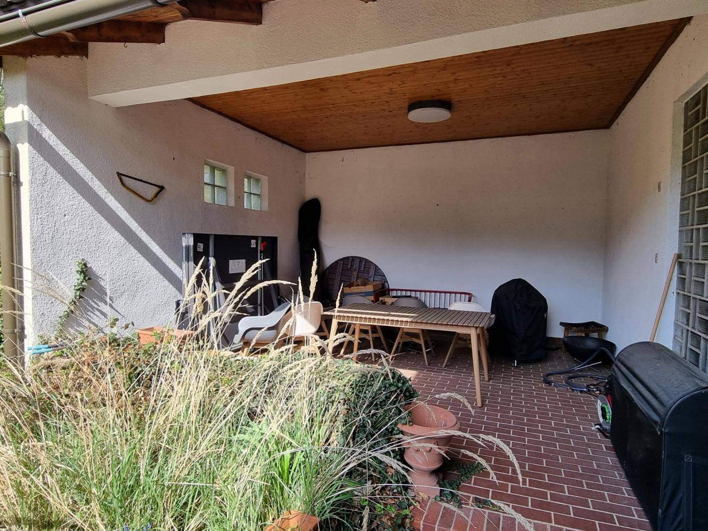 Überdachte Terrasse mit Ziegelboden und Gartenmöbeln, umgeben von hohen Gräsern.