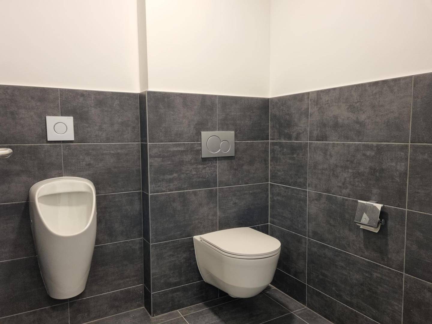 Modernes Badezimmer mit dunklen Fliesen, wandhängender Toilette und Urinal.
