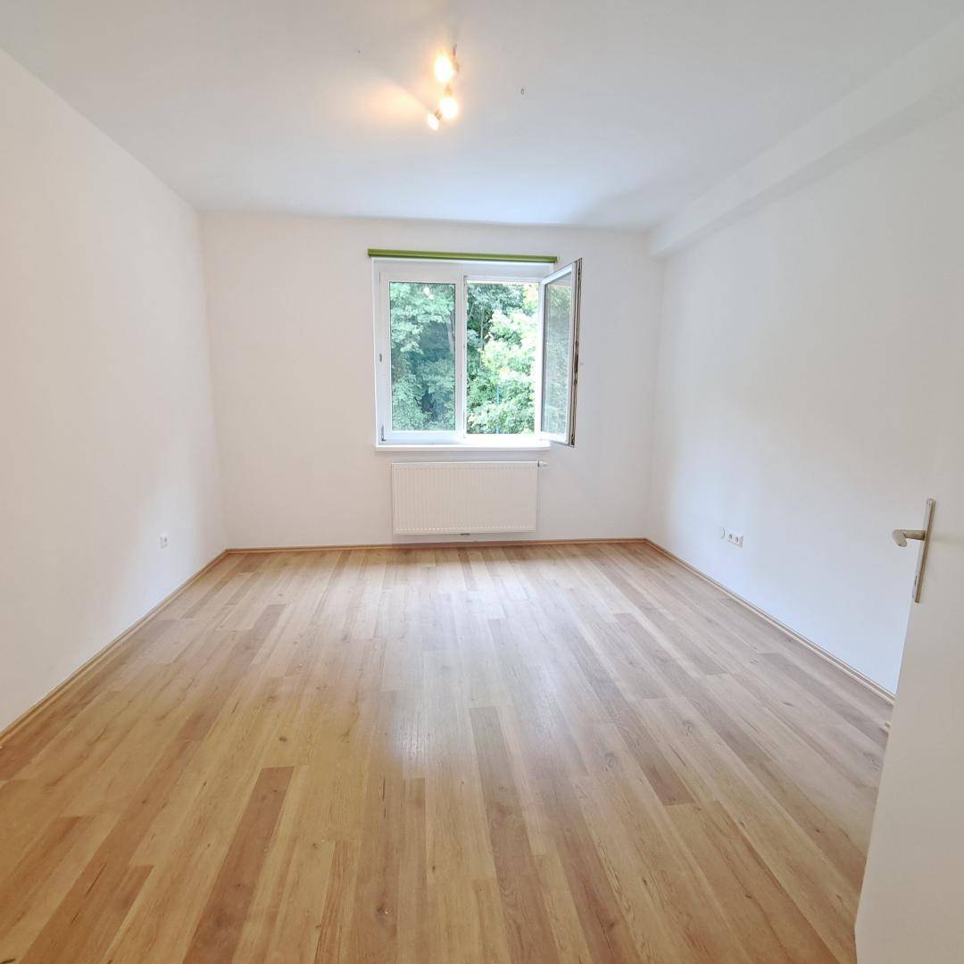 Helles Schlafzimmer mit Parkettboden und einem Fenster mit Blick ins Grüne.