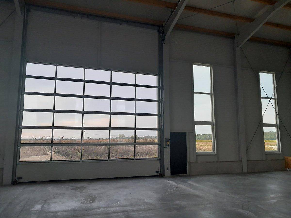 Heller Industrieraum mit großem Glasrolltor, hohen Fenstern und Blick auf die Landschaft.