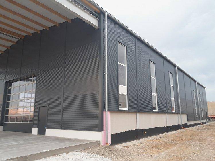 Seitenansicht des Industriegebäudes mit dunkelgrauer Wellblechfassade und hohen vertikalen Fenstern.