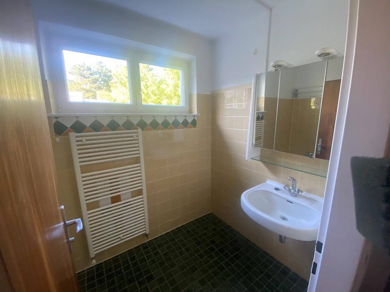 Badezimmer mit beigen Fliesen, Einzelwaschbecken, Spiegelschrank und einem Fenster für Tageslicht.