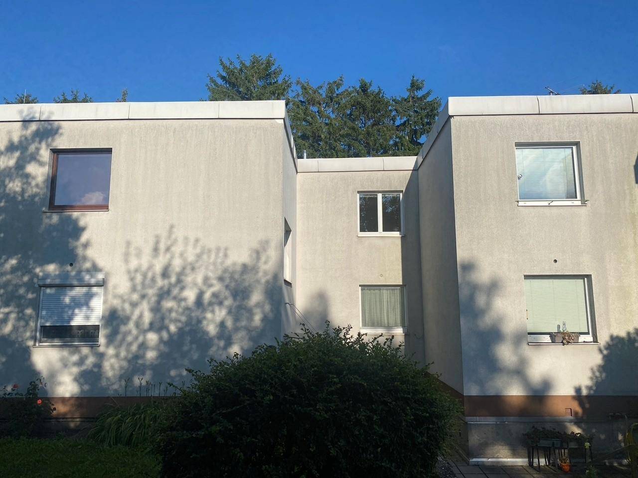 Schlichte Fassade eines Mehrfamilienhauses mit Flachdach und grüner Bepflanzung im Vordergrund.
