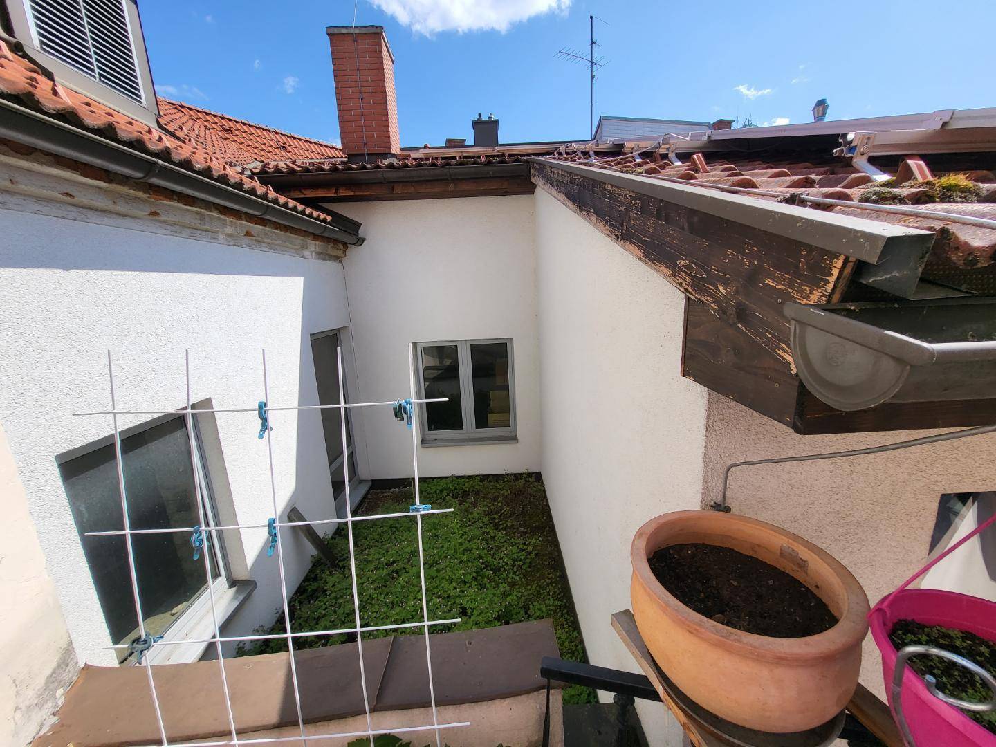 Blick von oben auf einen kleinen Innenhof mit grüner Bepflanzung und umliegenden Gebäudeteilen unter blauem Himmel.
