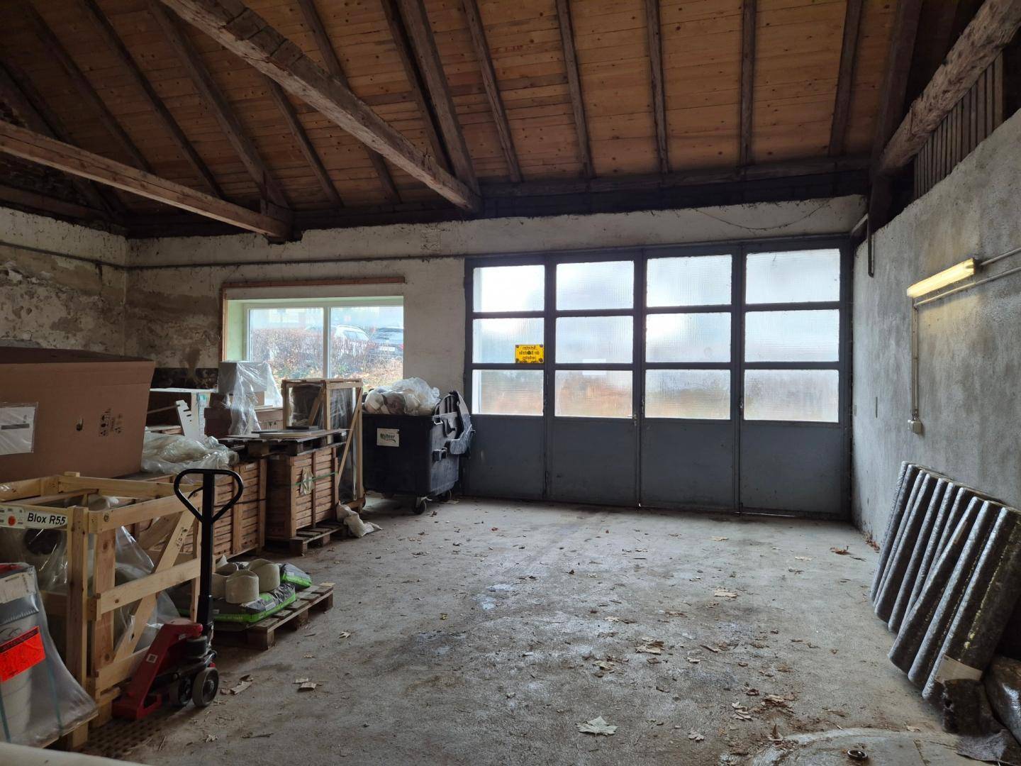 Großer Lagerraum mit Holzbalkendecke, Betonboden und einem Rolltor, gefüllt mit Baumaterialien.