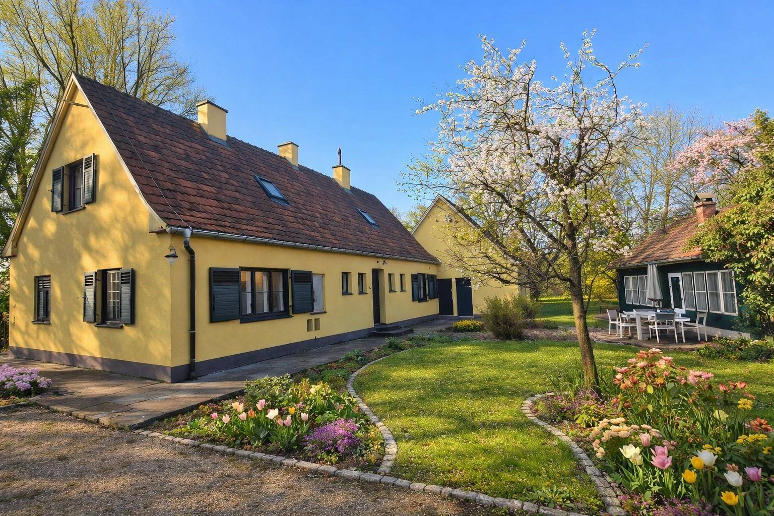 Charmantes gelbes Landhaus mit rotem Dach, umgeben von einem blühenden Garten und altem Baumbestand.