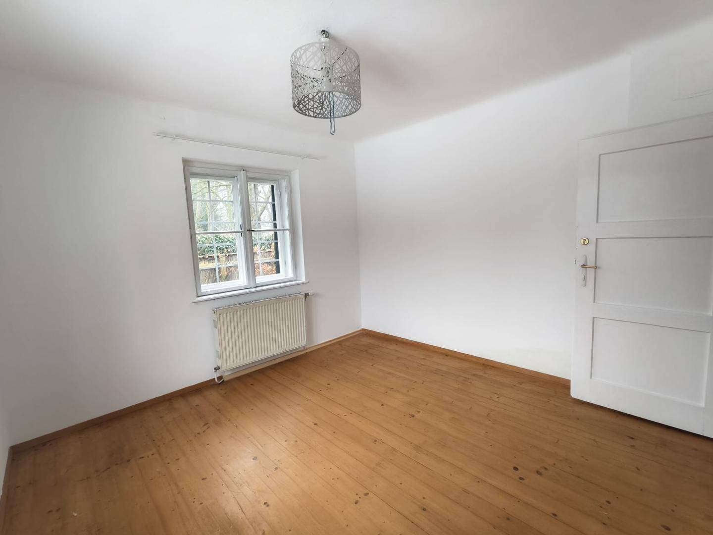 Kleineres Zimmer mit Holzboden, einem Fenster und einer schlichten Deckenleuchte.