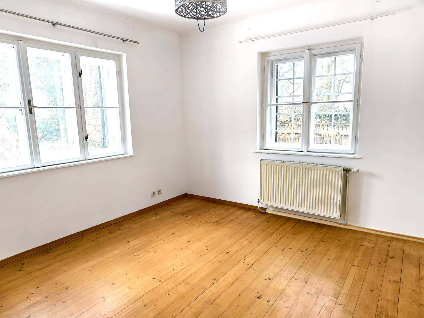 Helles Zimmer mit zwei Fenstern, Holzboden und einem Heizkörper unter dem Fenster.