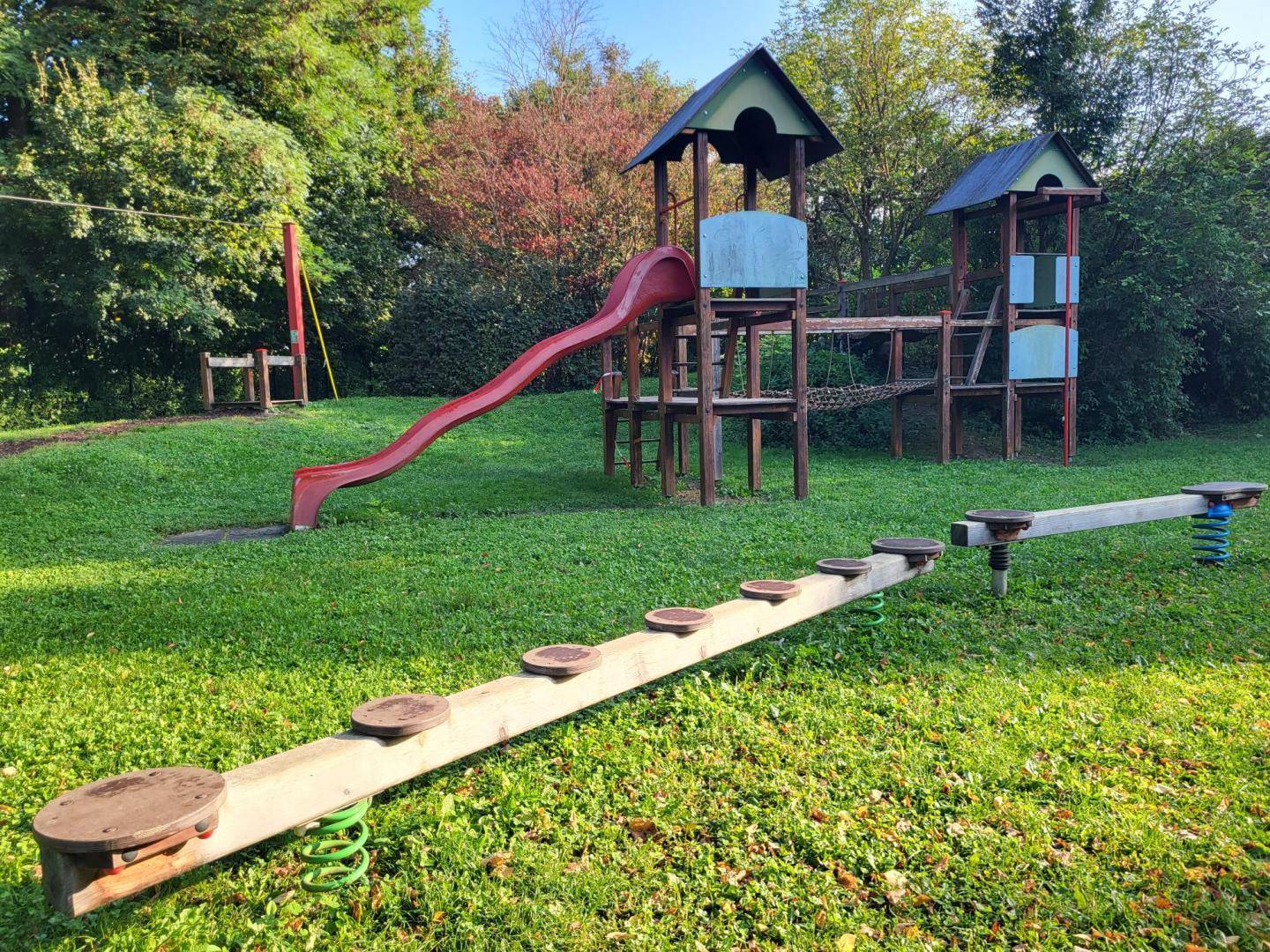 Kinderspielplatz mit Rutsche, Klettergerüst und Wippe auf einer grünen Wiese, umgeben von Bäumen.