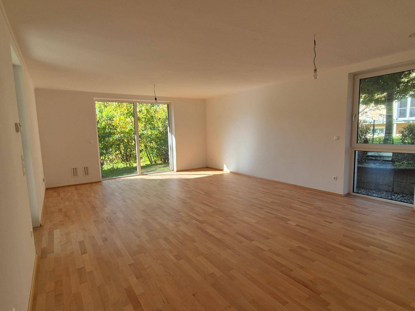 Helles, leeres Wohnzimmer mit hellem Parkettboden und großen Fenstern zum Garten.