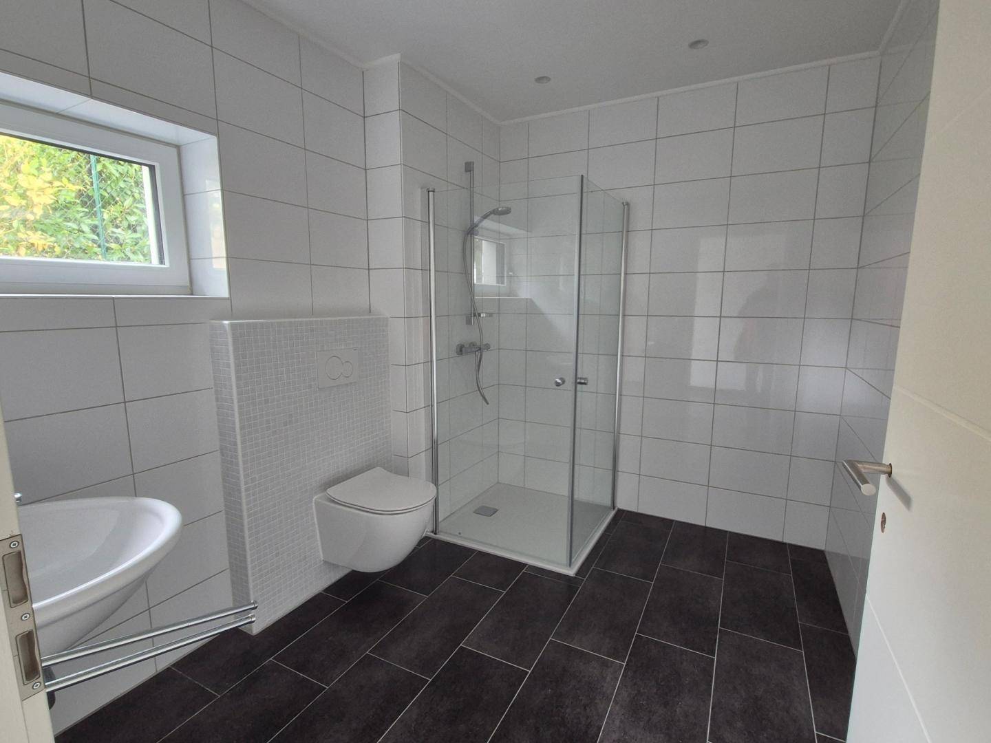 Modernes Badezimmer mit weißen Fliesen, ebenerdiger Dusche, wandhängendem WC und Fenster.