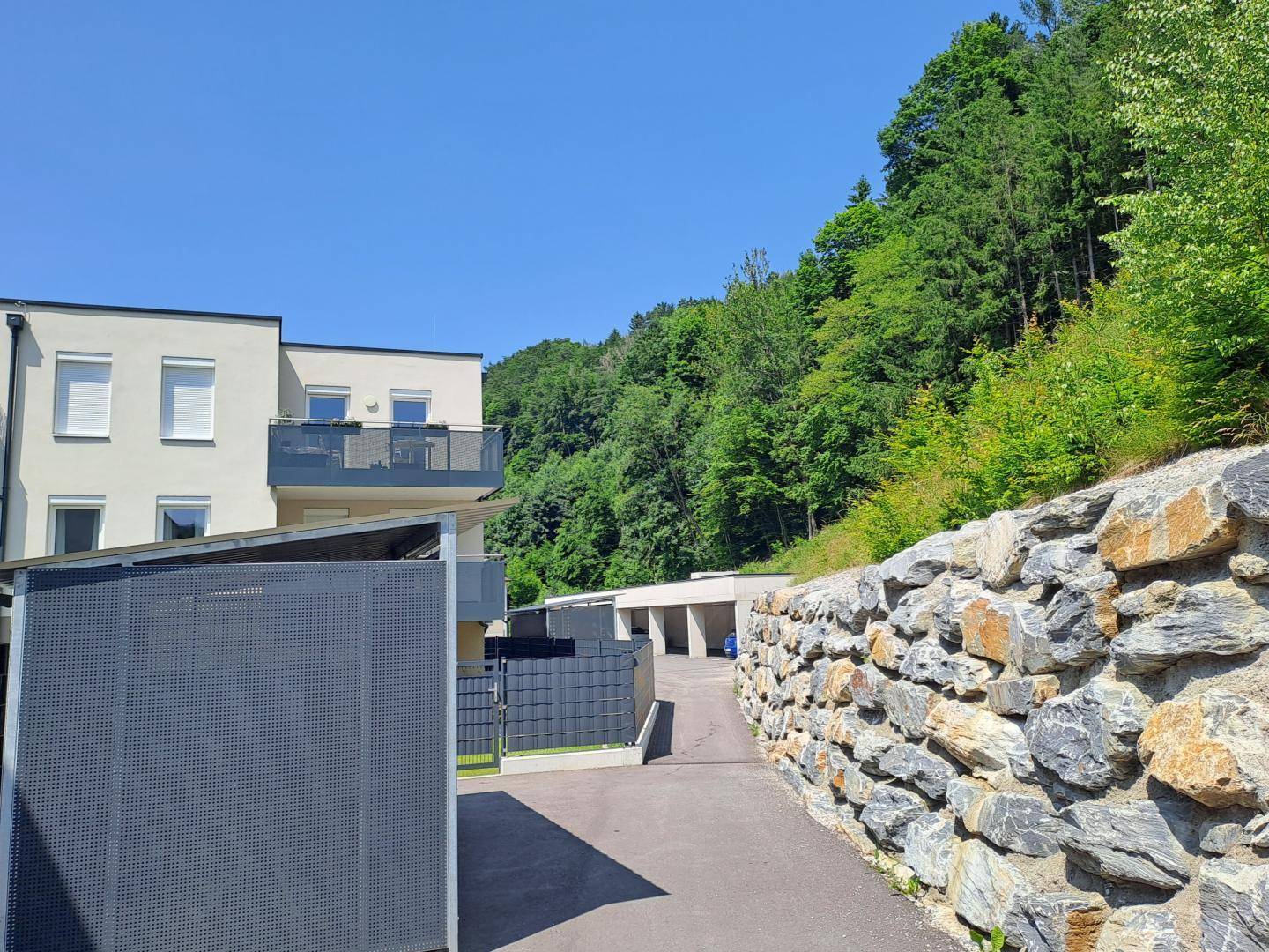 Moderne Wohngebäude am Hang mit Steinmauer und grüner, bewaldeter Umgebung.