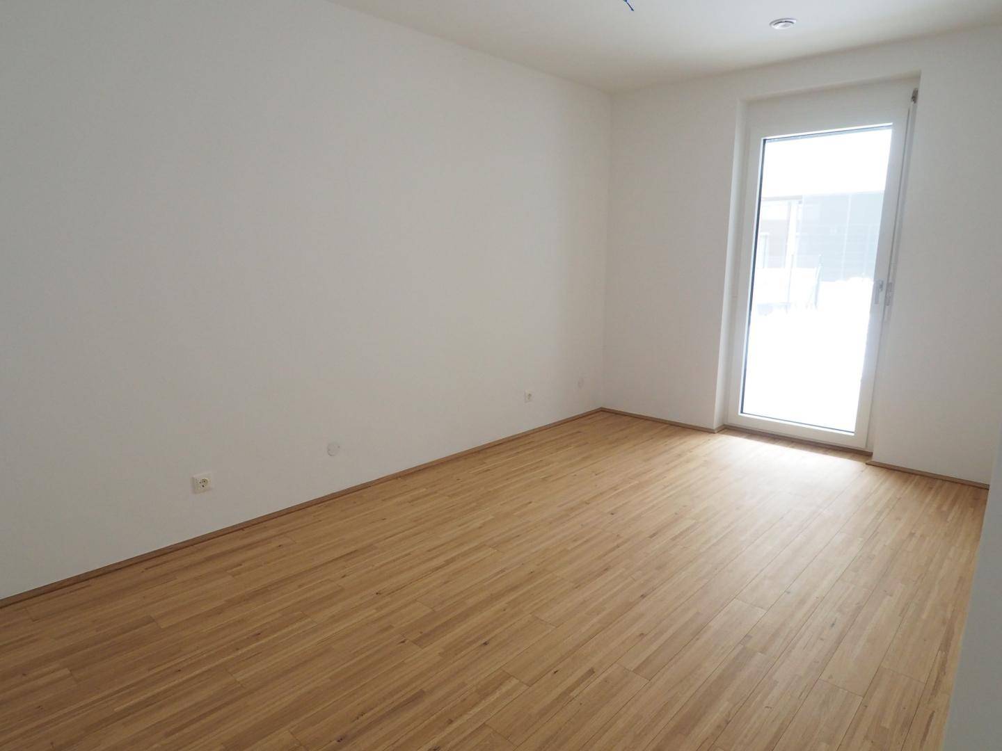 Helles Zimmer mit Holzboden und Zugang zu einem Außenbereich durch eine Glastür.
