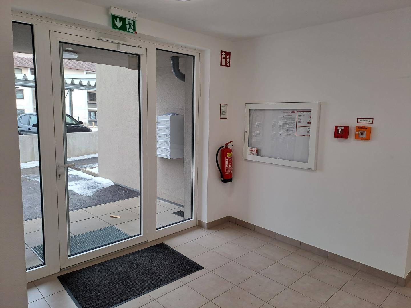 Heller Eingangsbereich mit Glastür, Fliesenboden, Feuerlöscher und Anschlagtafel in einem modernen Gebäude.