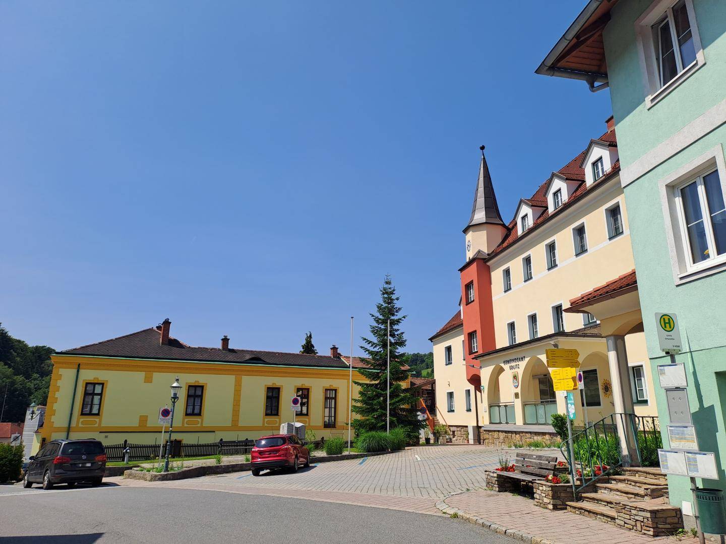 Dorfplatz mit traditionellen Gebäuden, einem Gasthof und einer Bushaltestelle.