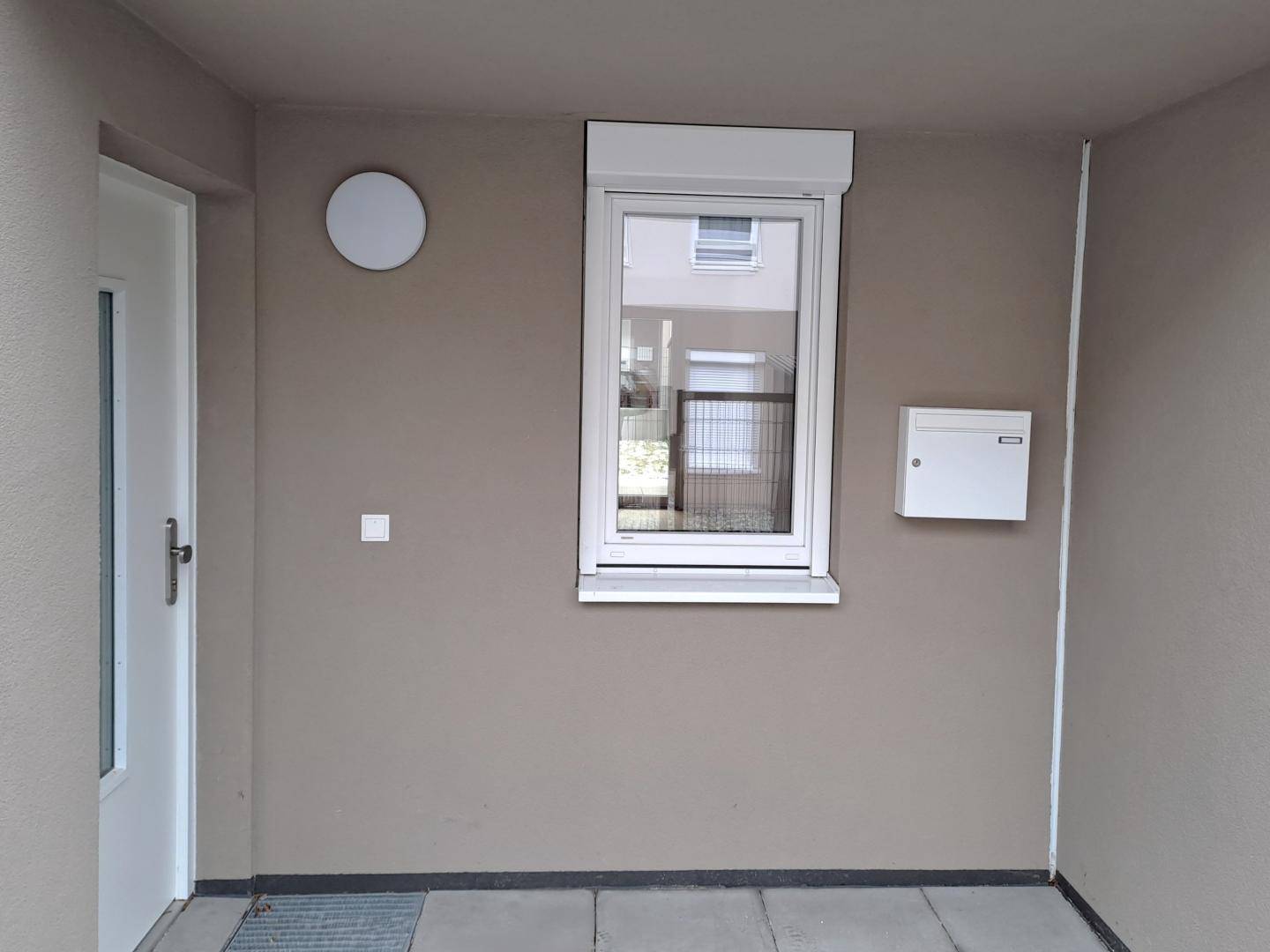 Überdachter Eingangsbereich mit Fenster, Briefkasten und schlichter Wandleuchte.