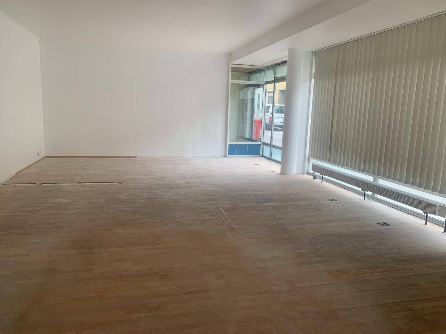 Geräumiger Innenraum mit Holzboden, weißen Wänden und großen Fenstern mit Jalousien.