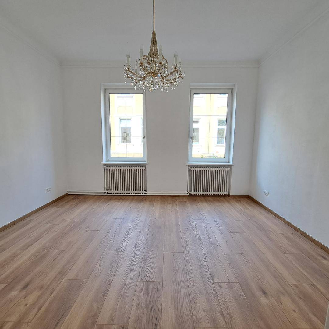 Helles, leeres Zimmer mit modernem Holzboden, hohen Decken und einem klassischen Kronleuchter.