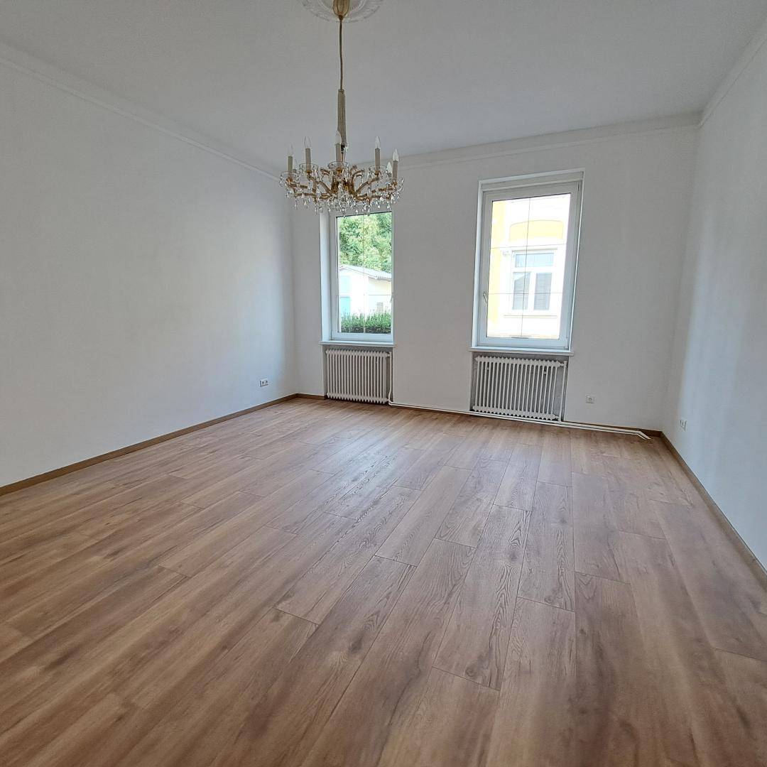 Geräumiges, helles Zimmer mit Holzboden, hohen Decken und einem eleganten Kronleuchter.