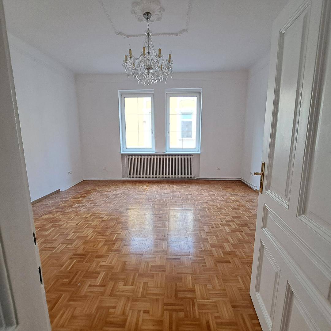 Geräumiges, leeres Zimmer mit klassischem Parkettboden, hohen Decken und einem eleganten Kronleuchter.