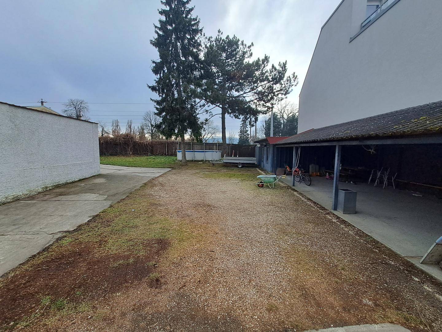 Weitläufiger Außenbereich mit Betonweg, Kiesfläche, überdachtem Unterstand und Bäumen im Hintergrund.