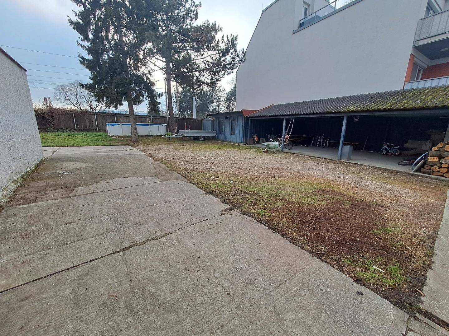 Großer Außenbereich mit Betonfläche, Kiesweg, überdachtem Stellplatz und kleinem Pool im Hintergrund.