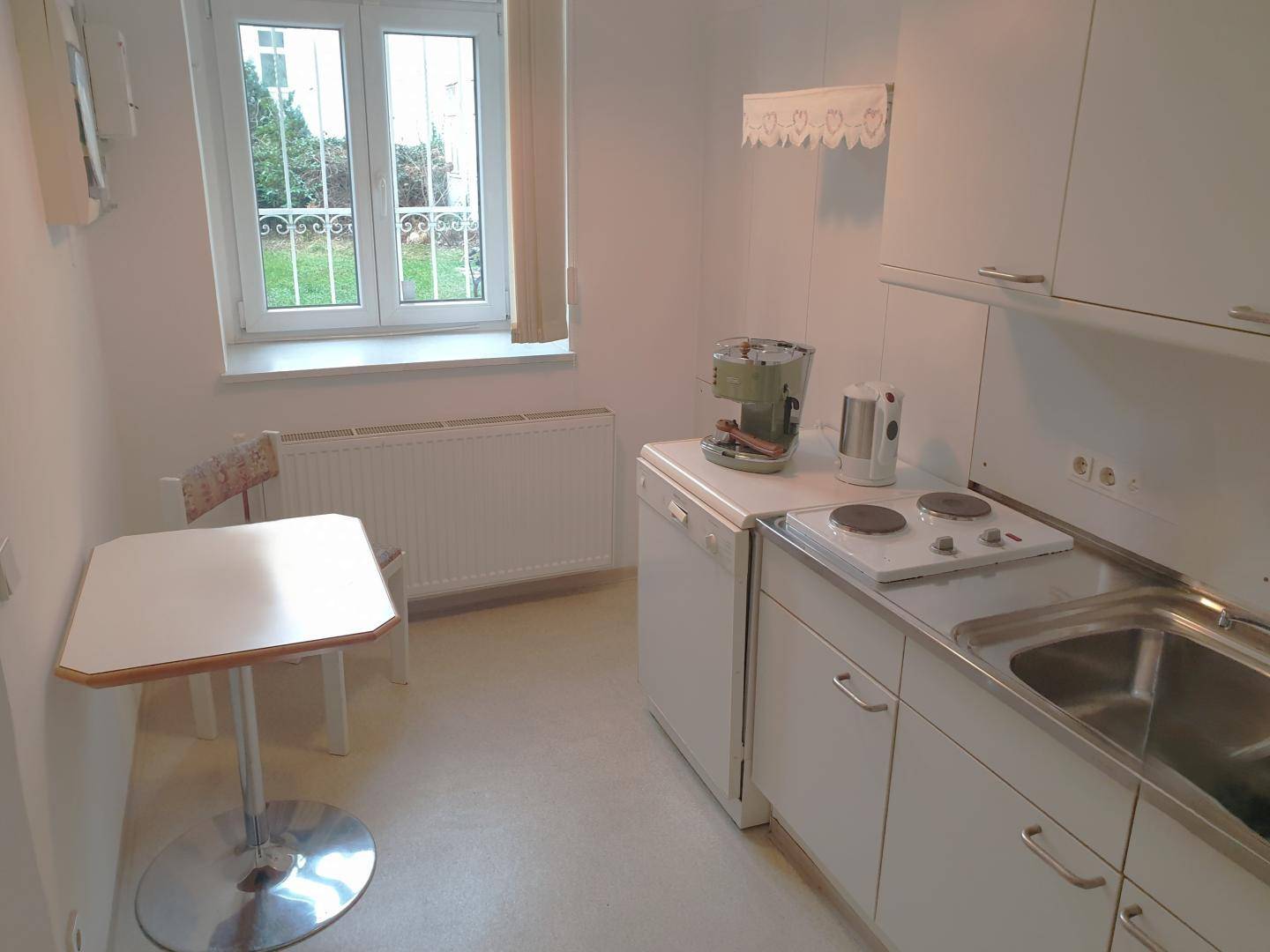Kleine Küchenzeile mit weißen Schränken, einem Fenster mit Blick ins Grüne und einem kleinen Esstisch.