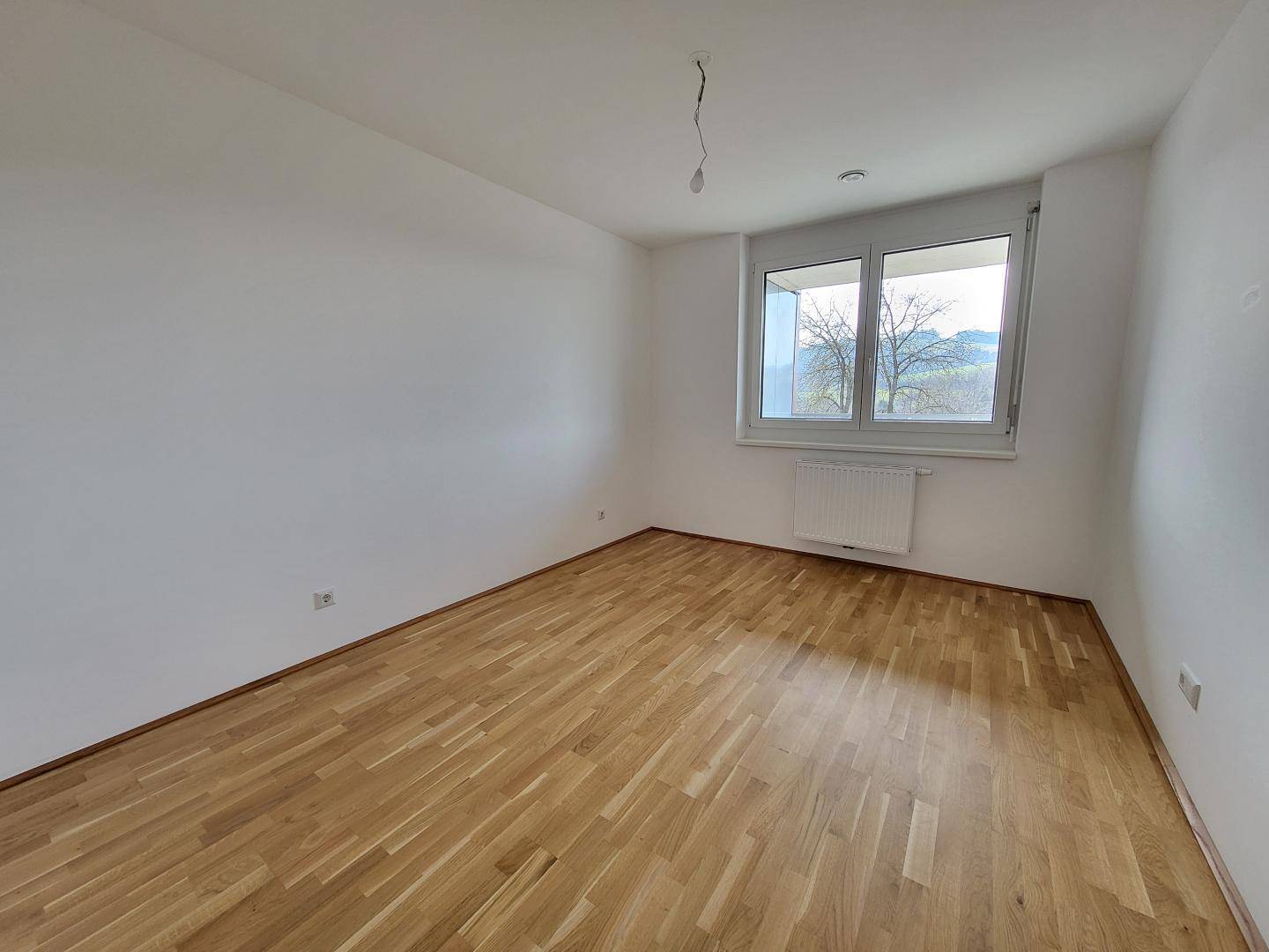 Schlafzimmer mit hellem Parkettboden und einem Fenster mit Blick ins Grüne.