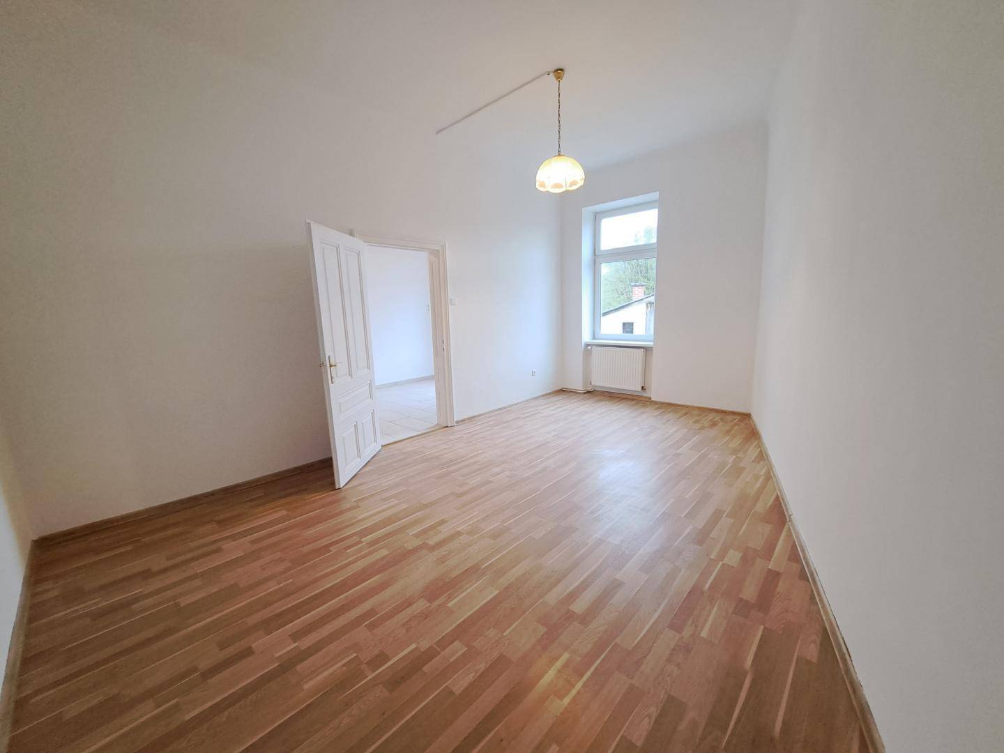 Leeres Zimmer mit Laminatboden, weiß gestrichenen Wänden und einem Fenster.