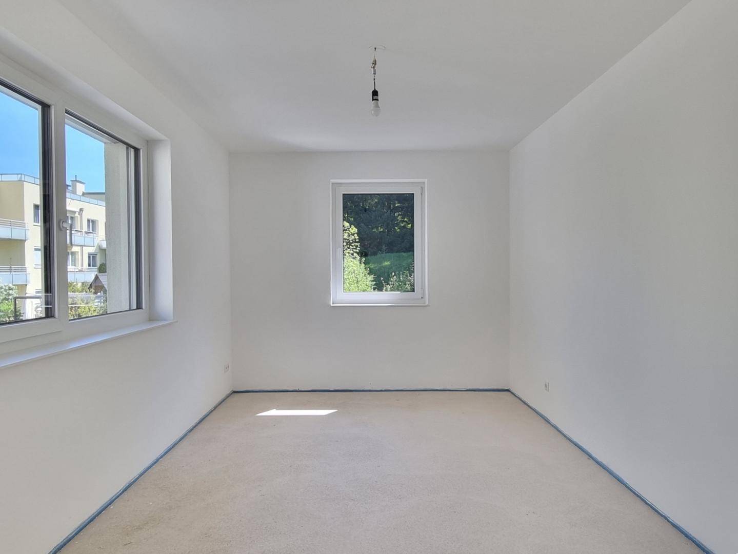 Unmöbliertes Schlafzimmer im Rohbau mit zwei Fenstern und Blick auf die Umgebung.