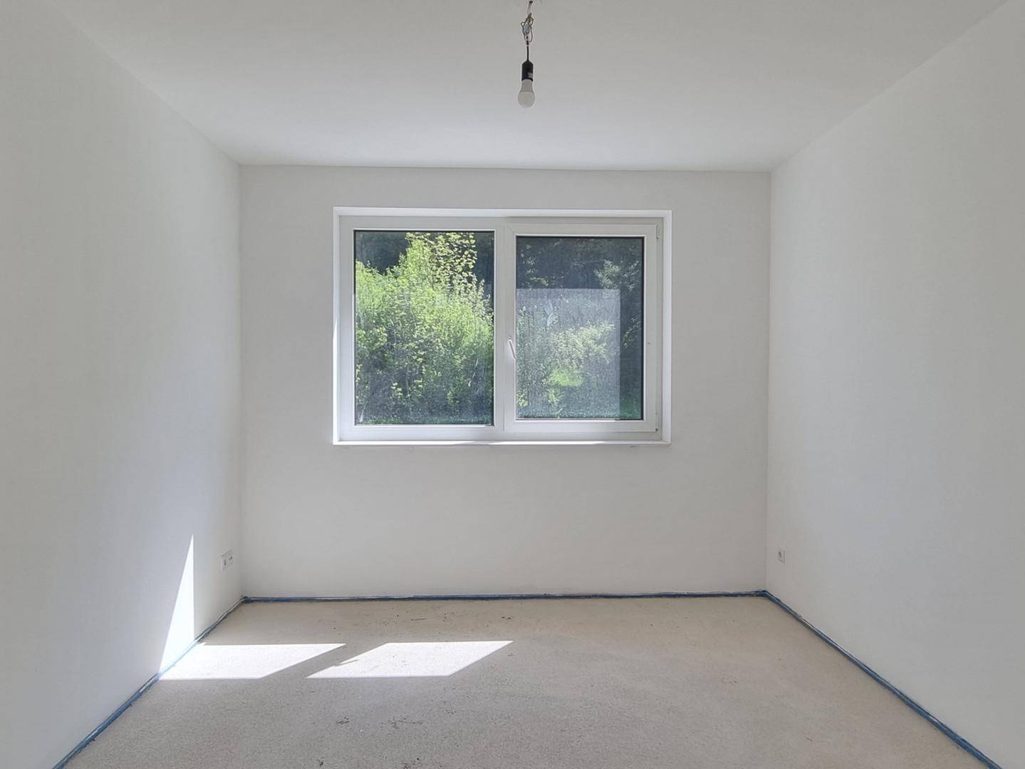Helles Schlafzimmer im Rohzustand mit großem Fenster und Blick auf die grüne Umgebung.
